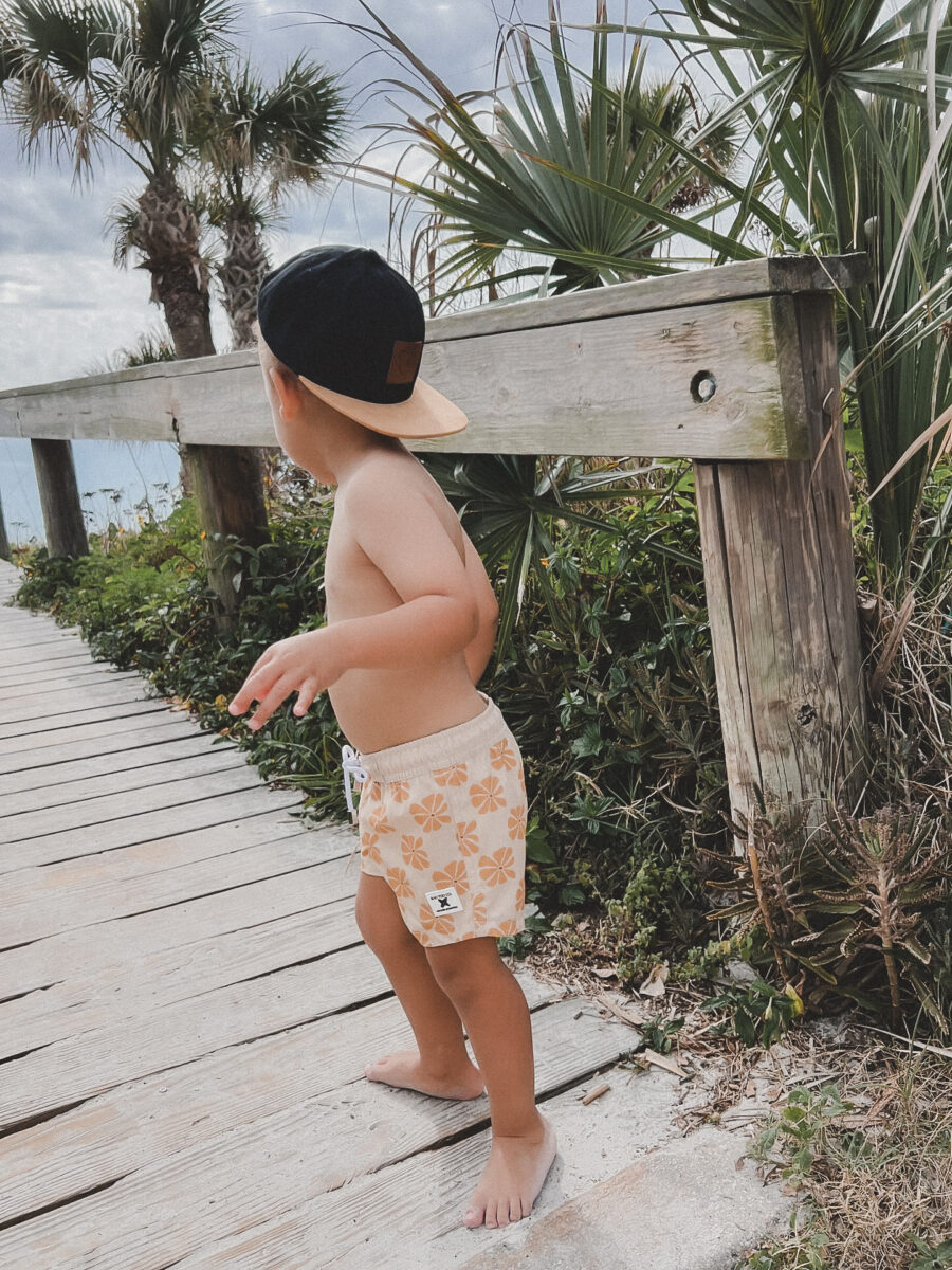 Boy wearing floral shorts by Rad Toddler stands on a pier by the beach