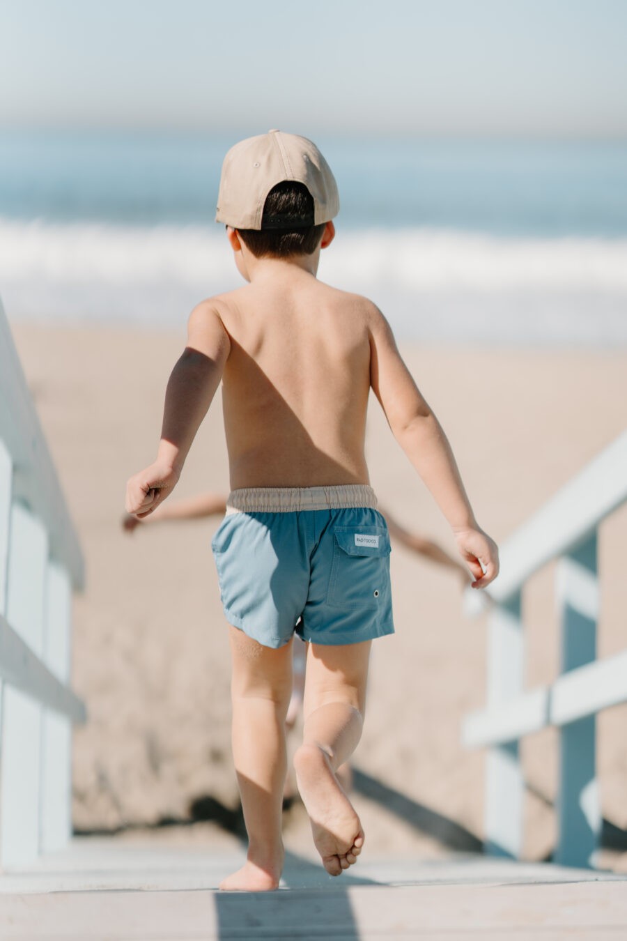 a boy is running down a lifeguard tower while wearing blue board shorts and beige hat by Rad Toddler