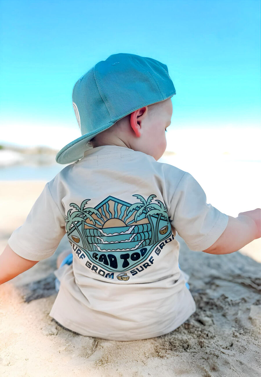 baby boy is sitting on the beach playing with the sand while wearing his surf grom tee by Rad Toddler