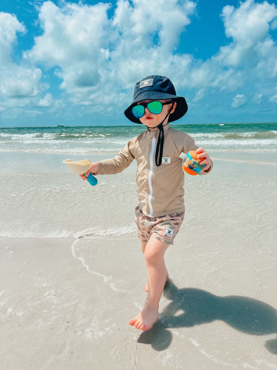 Boy playing by the ocean wearing a protective bucket hat, glasses, rash guard shirt, and board shorts by Rad Toddler