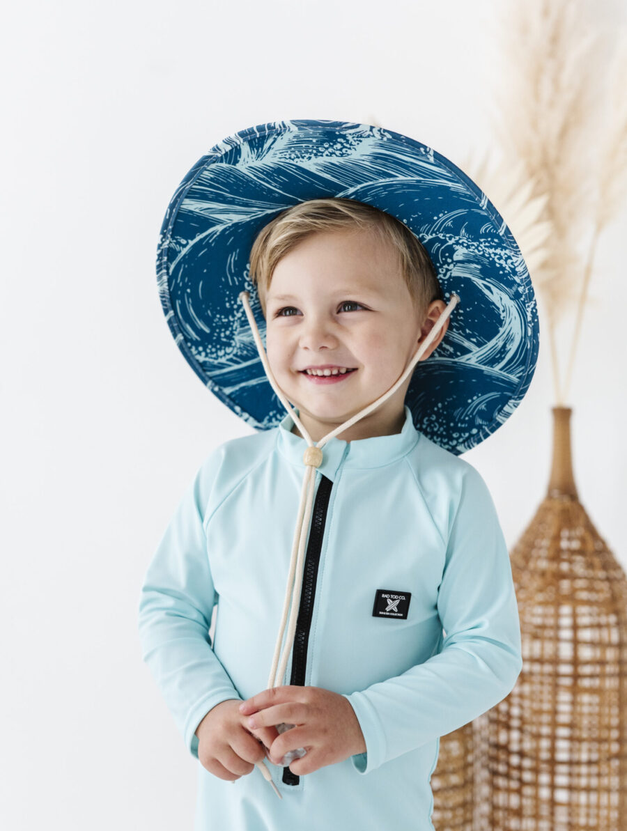 a boy smiles and looks on happily as he wears his straw hat and blue rash guard by Rad Toddler